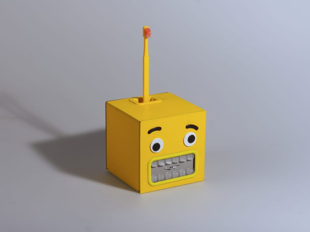 The BrushBuddy cube at a three-quarter angle, the toothbrush standing in its top dock, the worried monster face turned to camera, on a clean gray field.