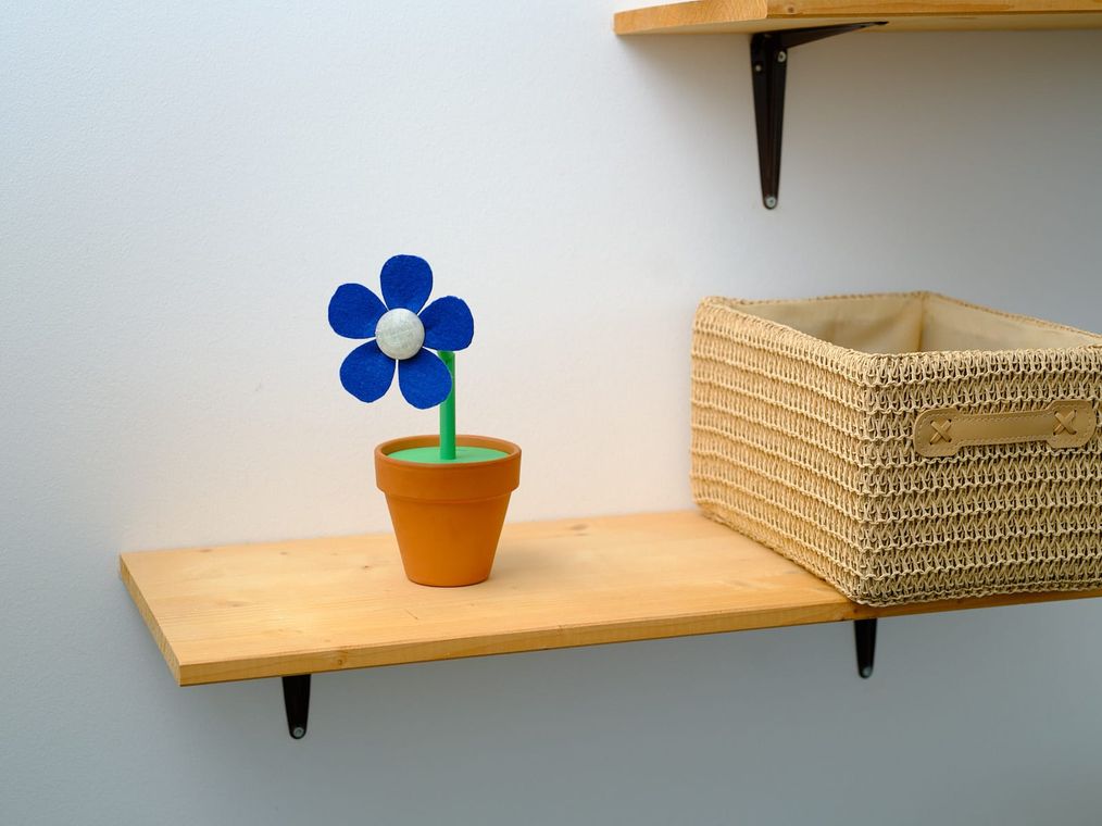 DataBloom on a wooden wall shelf beside a woven basket, its stem bent partway down, in a home interior.