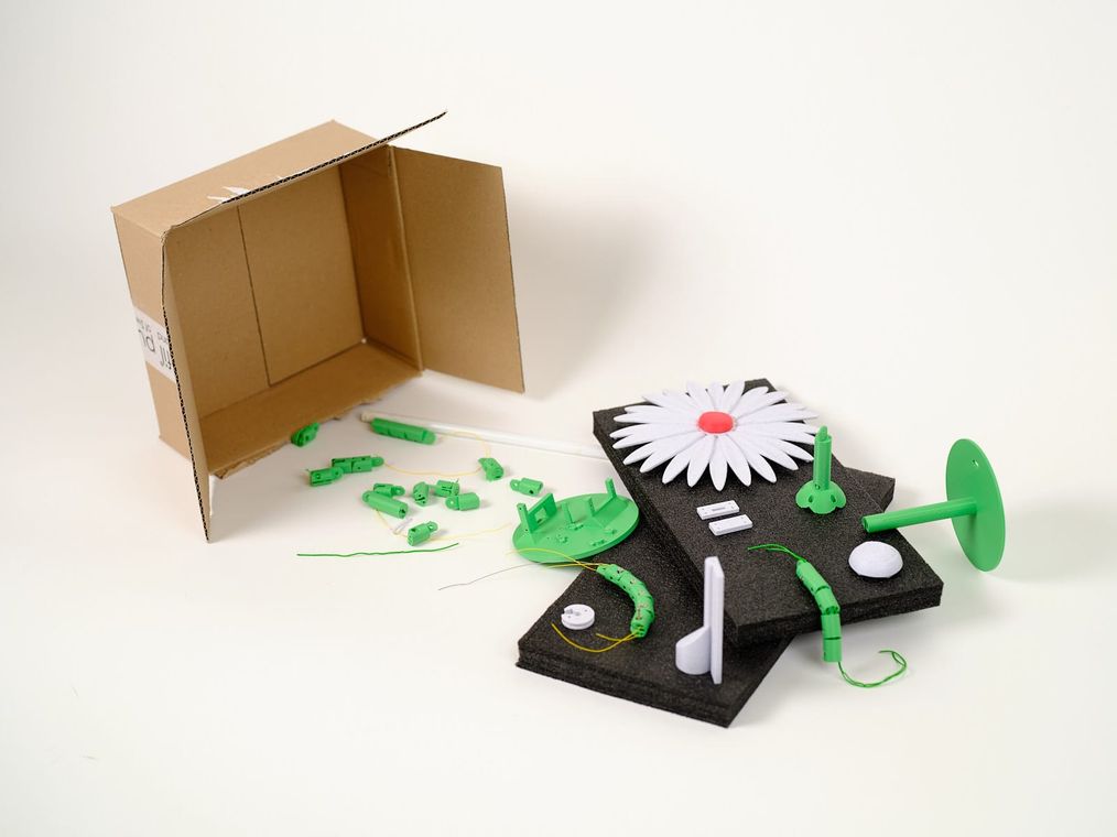 Printed parts from across the project laid out on foam beside a shipping box: the white low-fidelity daisy alongside the final green stem links, servo arm and electronics.