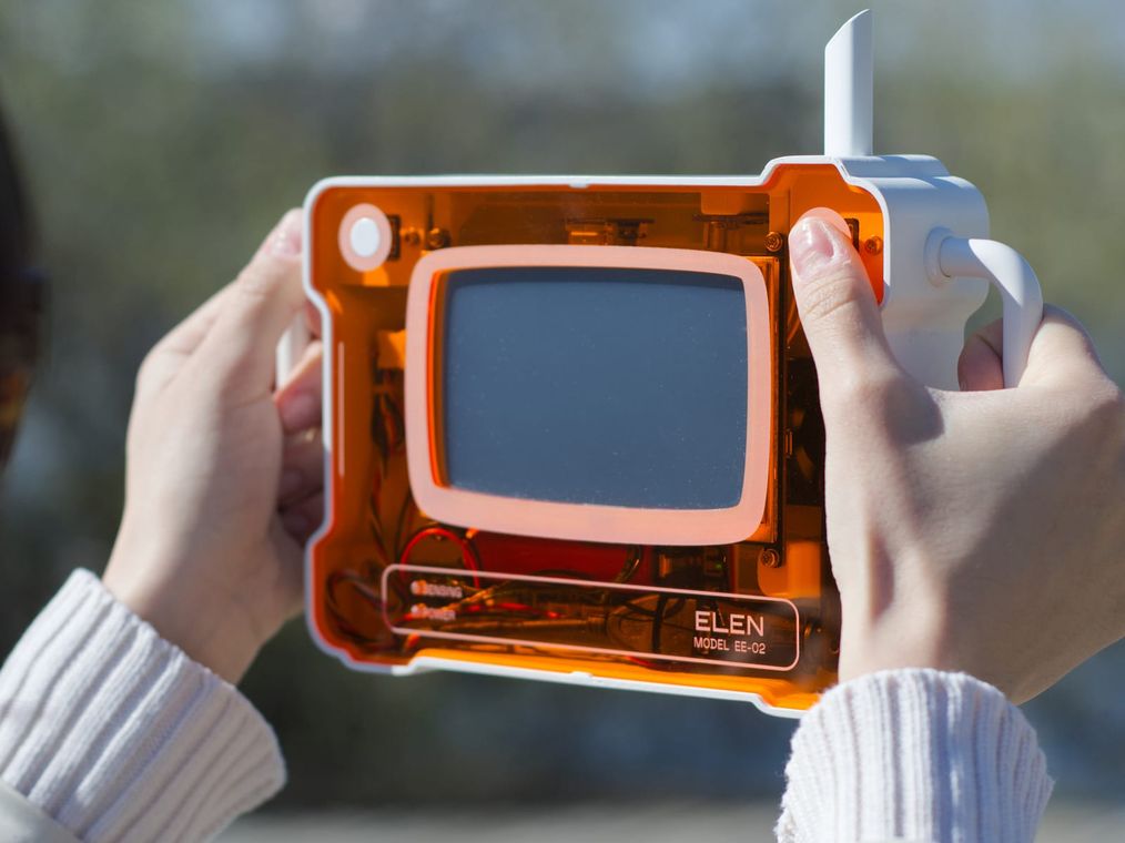 A person holds ELEN up in both hands outdoors, framing the space through its screen, the translucent orange body lit by daylight.