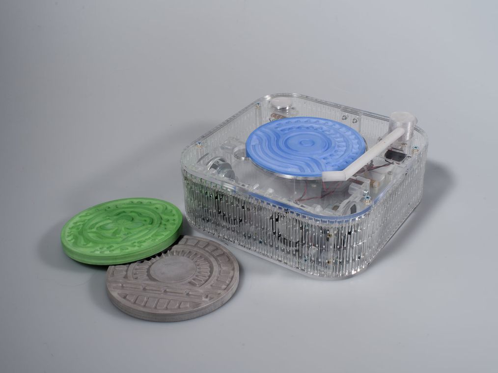 The transparent acrylic listening box with its tonearm, seen three-quarter, beside the three plaster disks.