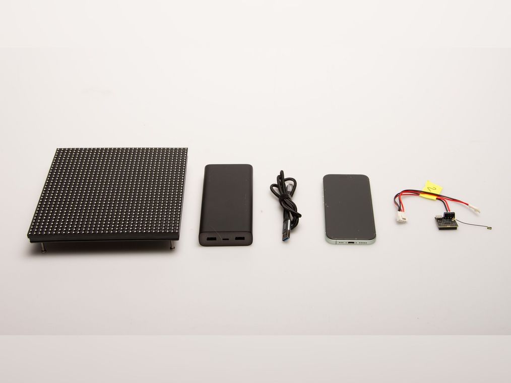 The build laid out on a light surface: the 32×32 LED matrix panel, a power bank, a USB cable, a smartphone, and the small ESP32 board with its antenna and battery harness.