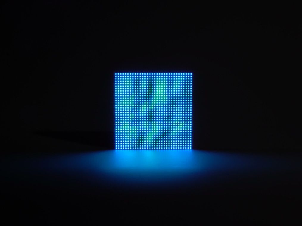 A single Meeting Pond unit standing in a dark room, glowing a calm even blue across its 32×32 LED grid, its light pooling on the surface below.