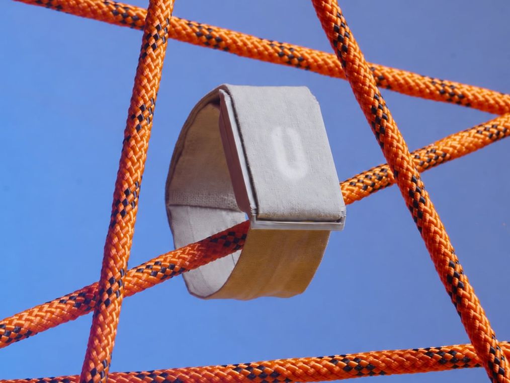 The wearable suspended in a lattice of orange cords against a cobalt sky, its oblong gauge glowing.