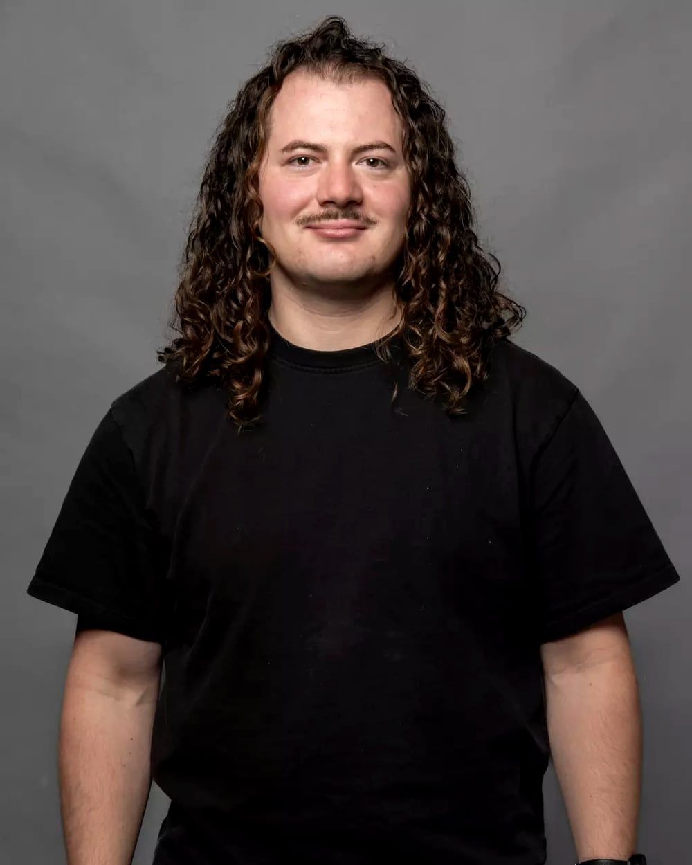 Studio portrait of Jérémy Martin, shoulder-length curly hair and a moustache, wearing a black t-shirt against a gray backdrop.
