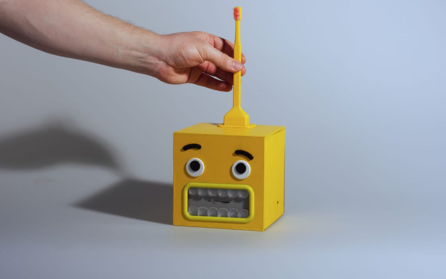 A hand lifts the toothbrush from the dock on top of the BrushBuddy cube, which has a worried yellow monster face.
