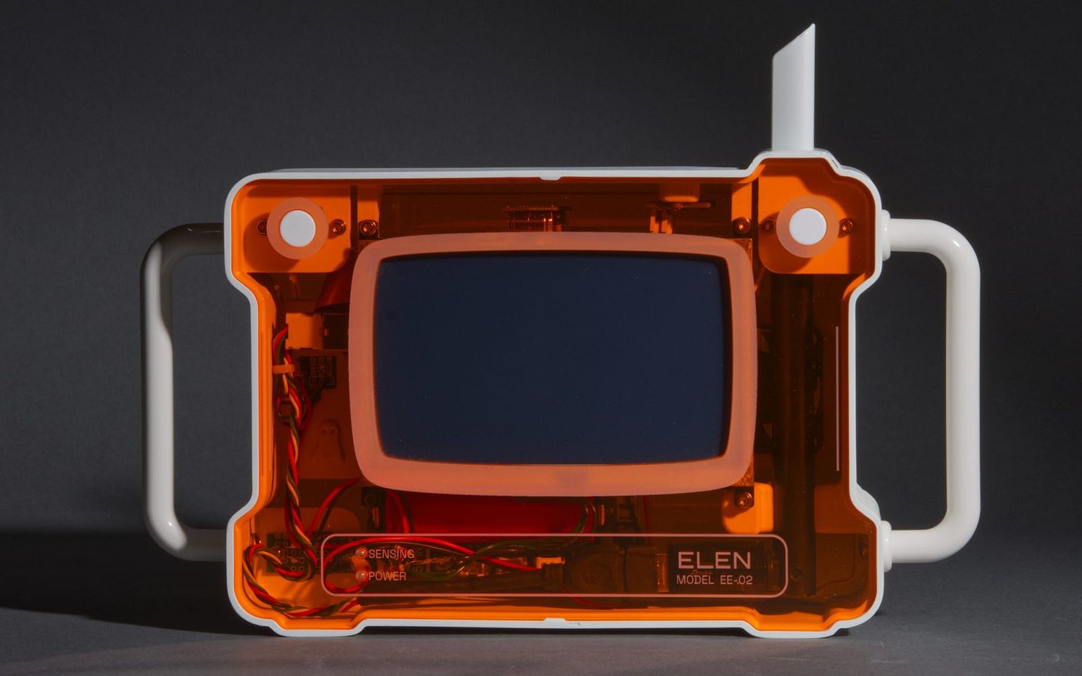 ELEN, a handheld camera with a translucent orange body and twin white handles, lit on a gray studio backdrop, its deep-blue screen showing through the plexiglass.