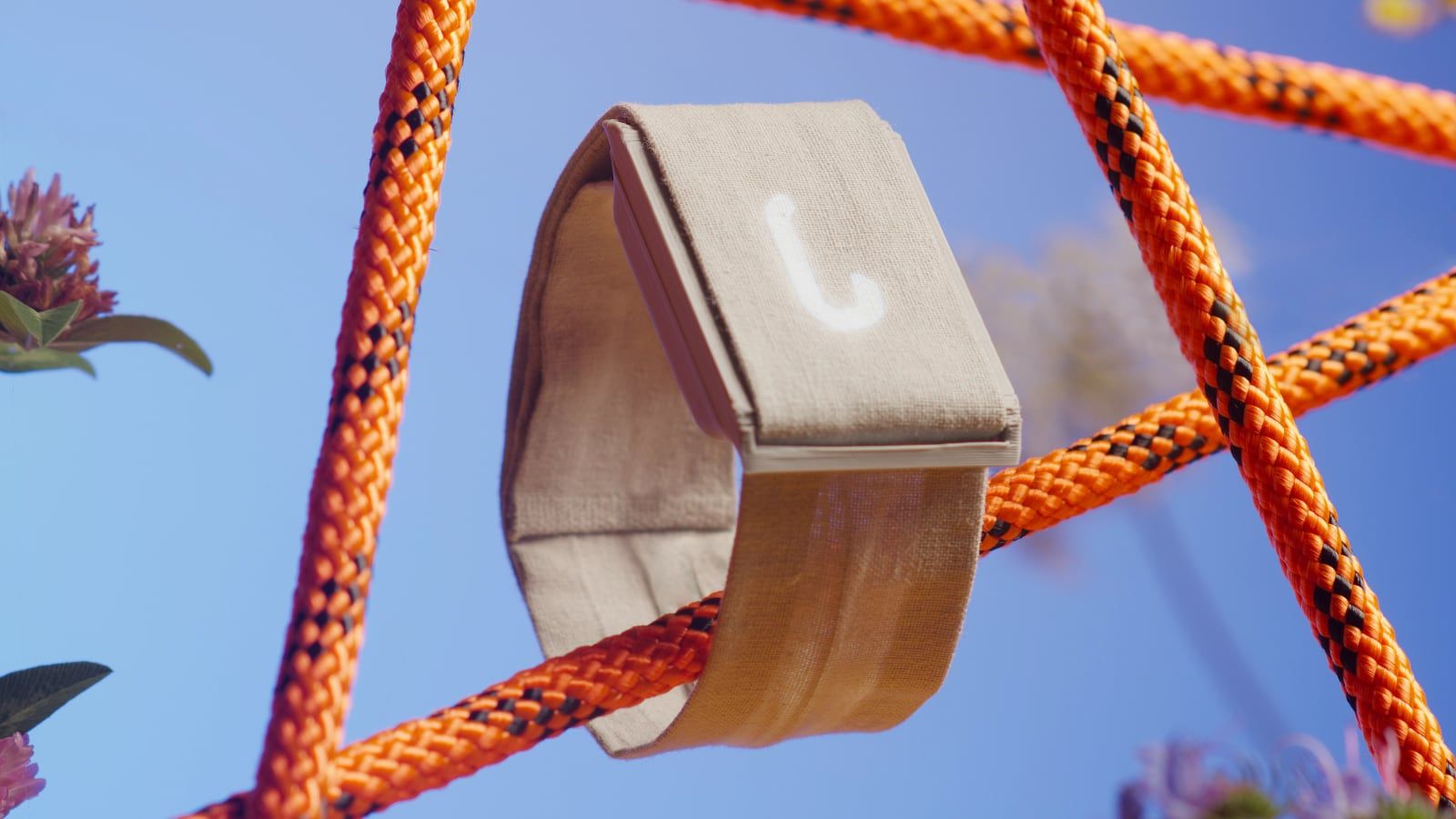 Thea featured card: the pale wrist cuff suspended in a lattice of orange cords against a cobalt sky with a sprig of wildflower, its ring gauge glowing.