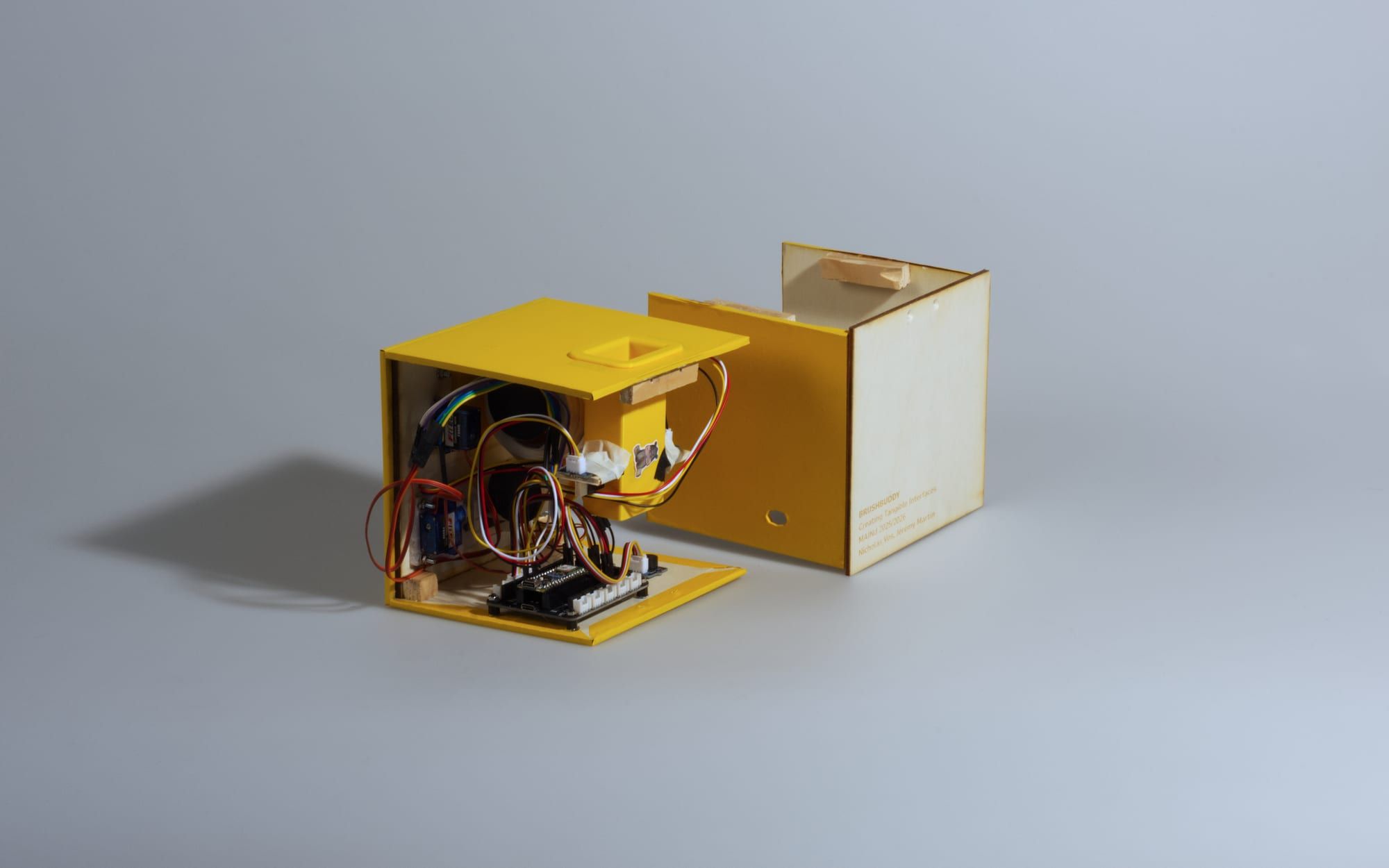 The cube opened into two halves: one holds the ESP32 controller, three servos, the dock sensor, a buzzer and wiring; the other is the plywood shell laser-etched with the BrushBuddy name and course credit.