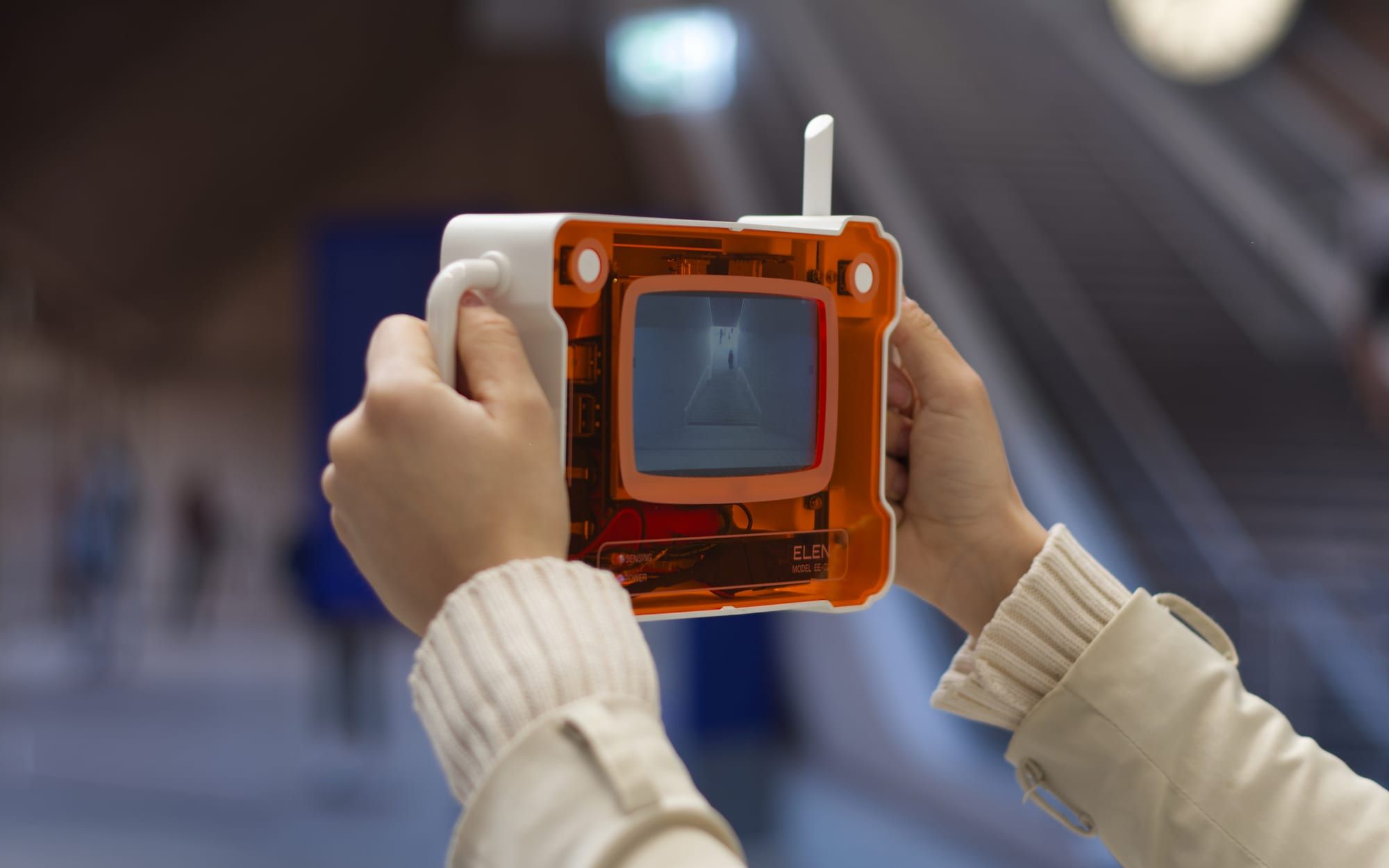 A visitor holds ELEN up in both hands in an indoor public space, framing the room through its screen, the orange body glowing against the daylight behind.