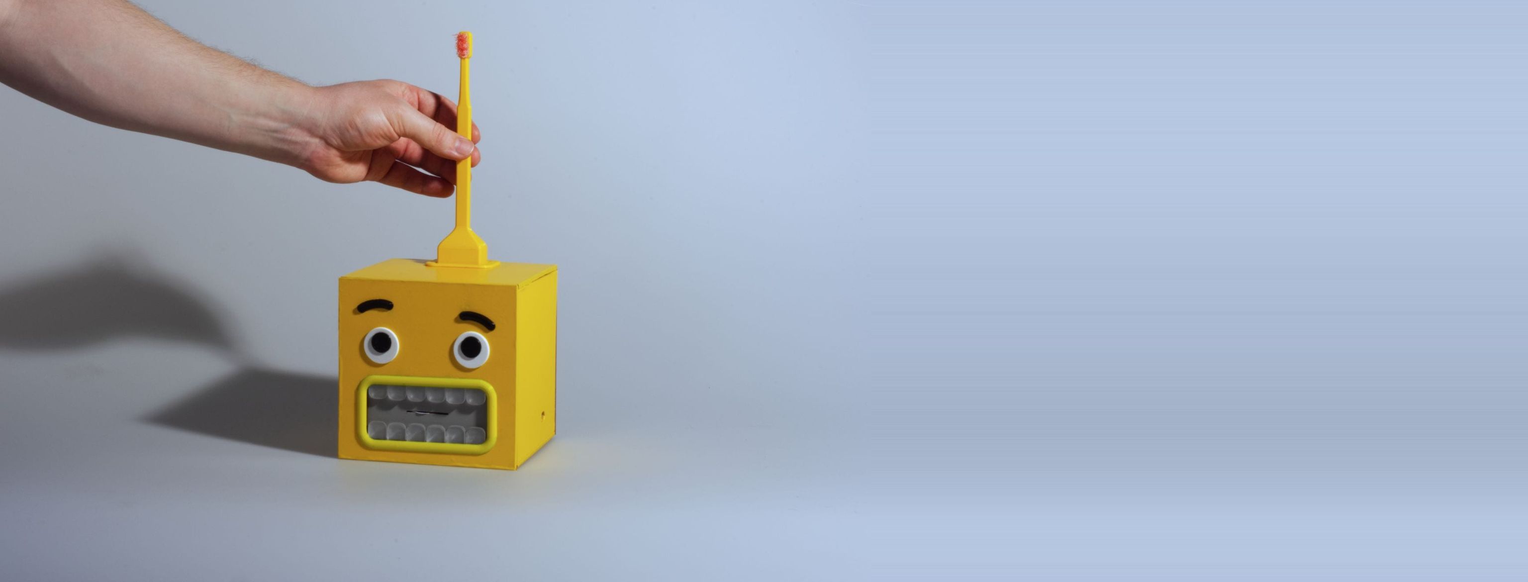 A hand lifts the yellow toothbrush from the dock on top of the BrushBuddy cube, whose worried monster face — raised brows, googly eyes, a mouth of twelve gray teeth — waits below on a wide gray field.