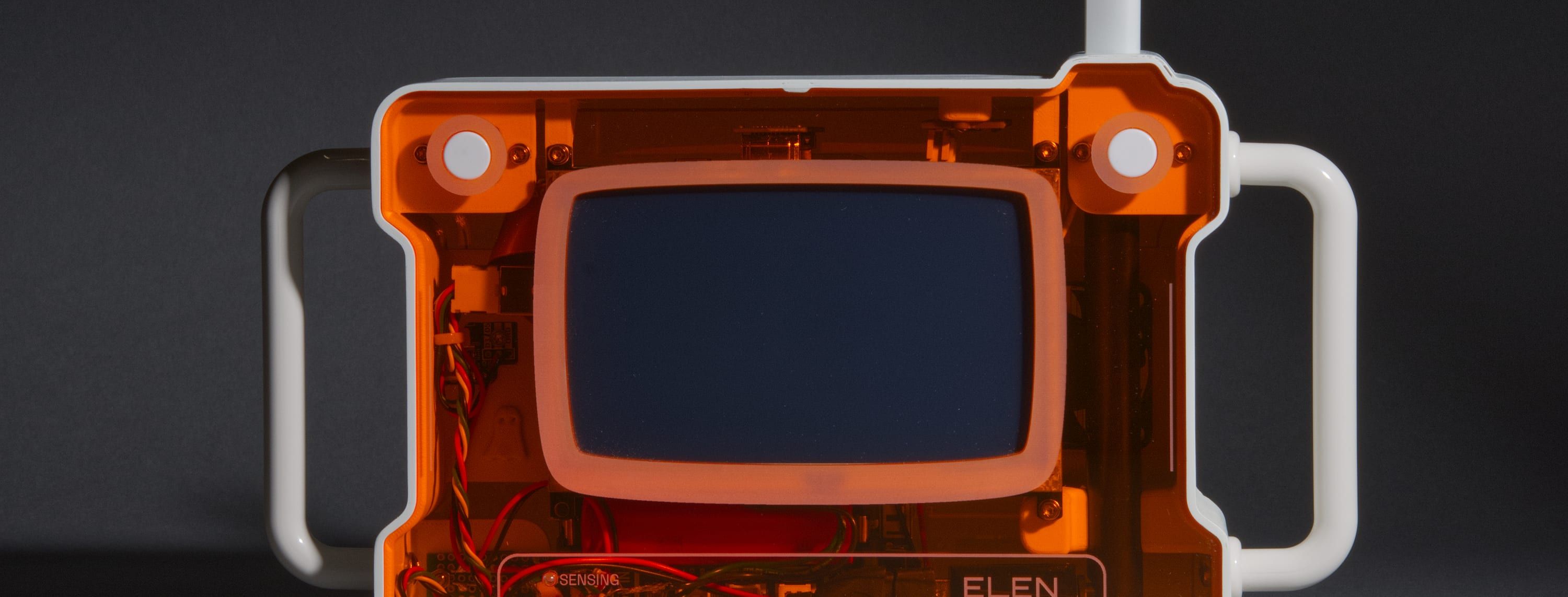 ELEN, a handheld camera with a translucent orange 3D-printed body and twin white handles, lit on a gray studio backdrop. Its deep-blue screen and internal wiring show through the plexiglass.