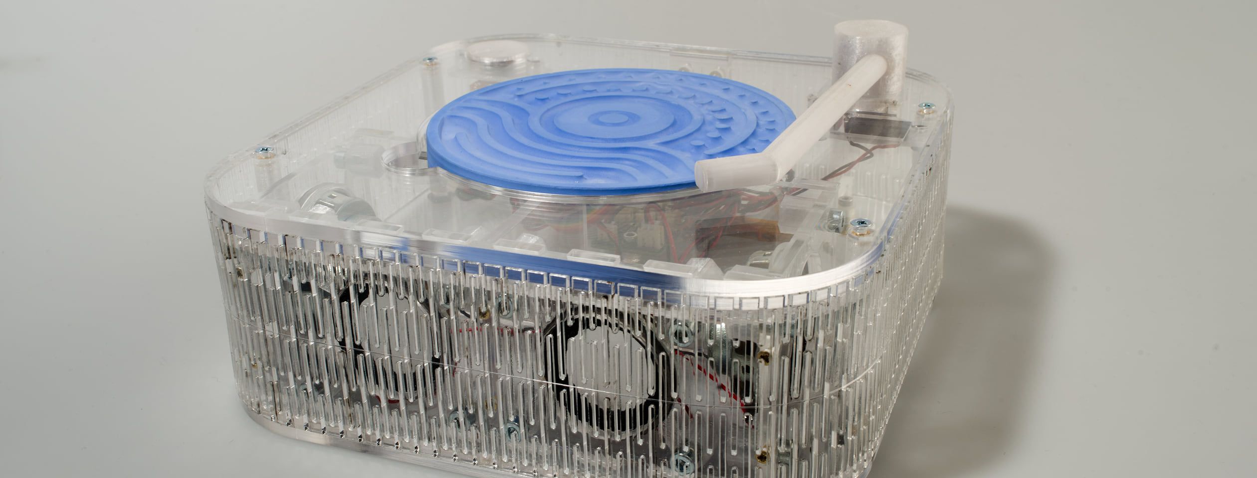 The Human Loci listening box: a transparent acrylic box with internal speakers and a white tonearm, a textured blue plaster disk resting on its deck, on a seamless gray backdrop.