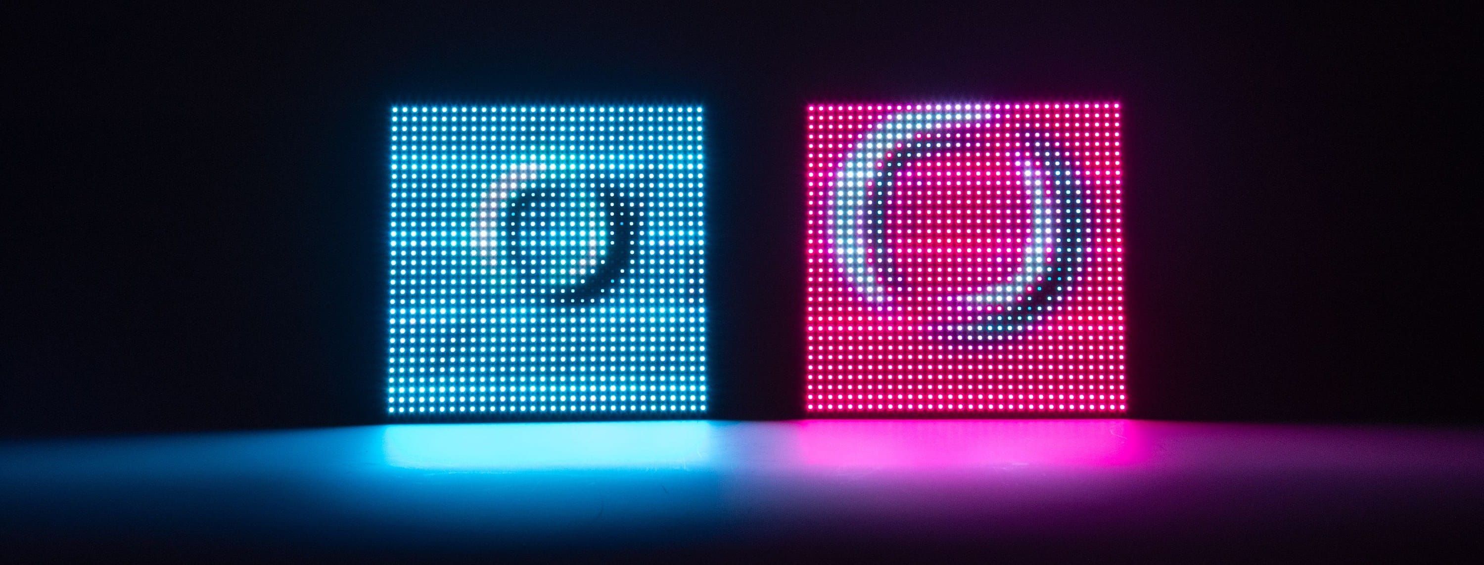 Two Meeting Pond units standing side by side on a reflective surface in a dark room, one glowing blue and the other pink, each showing a circular ripple across its 32×32 LED grid, their light pooling below.