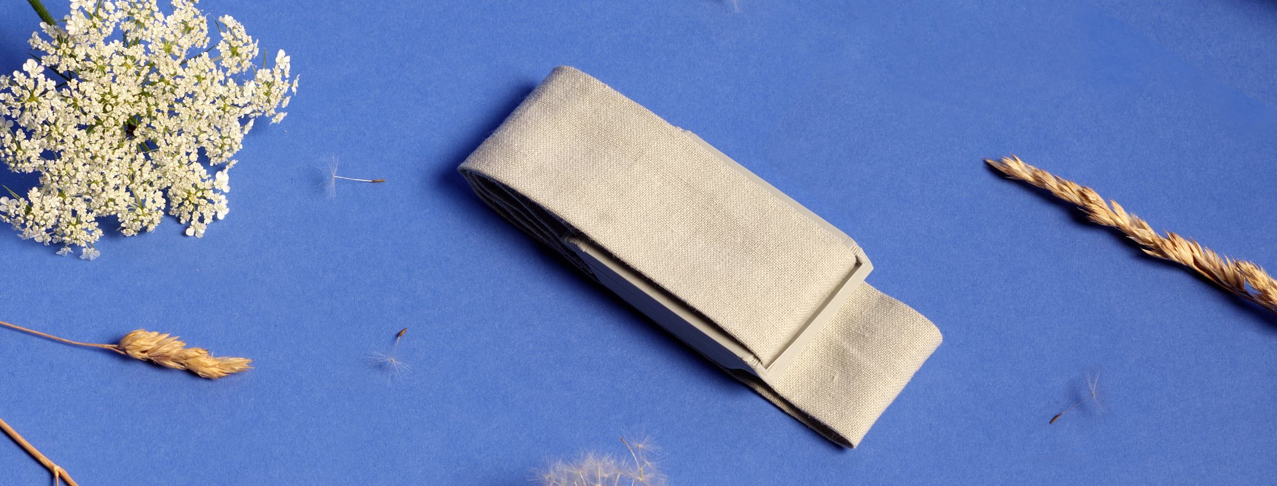 A top-down view of the Thea wearable, a pale housing on a fabric band, resting on a deep cobalt surface scattered with dried wildflowers, grasses, and seed heads.