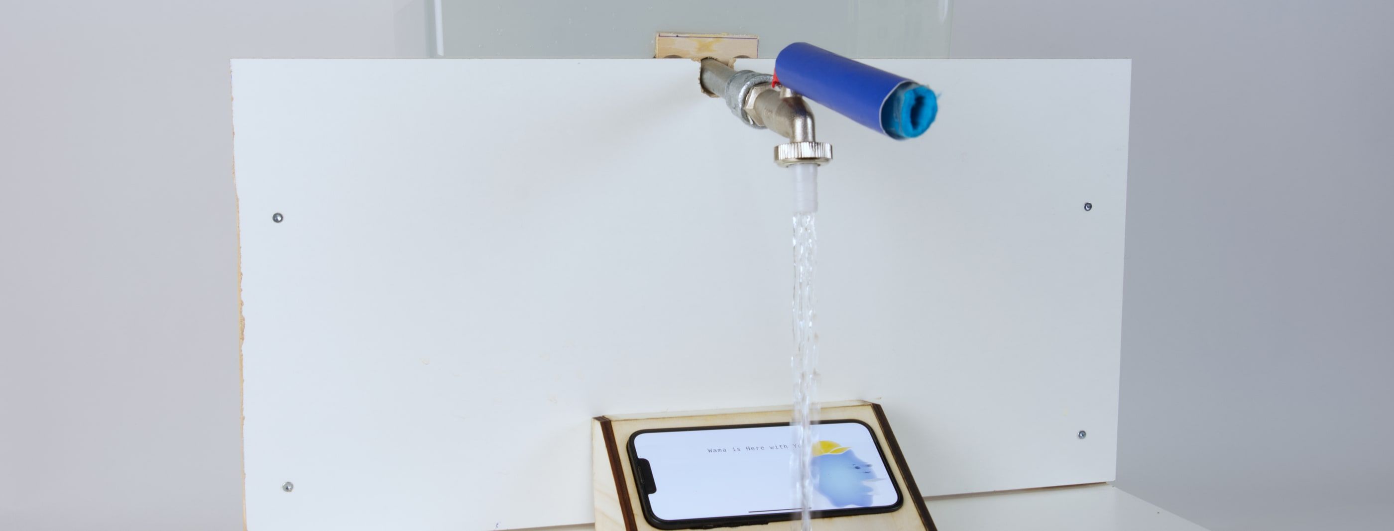 Wama running on a phone in a laser-cut wooden holder at a built sink rig: a tap pours into a clear basin while the screen shows a small blue cloud character.